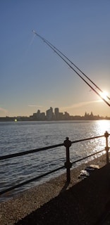 The image captures a scenic view of a waterfront during sunset. A sleek fishing rod is prominently angled in the foreground against the backdrop of a calm water surface reflecting the sun's golden rays. On the horizon, a city skyline with various buildings is silhouetted against the clear sky.