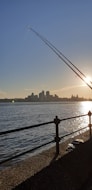 The image captures a scenic view of a waterfront during sunset. A sleek fishing rod is prominently angled in the foreground against the backdrop of a calm water surface reflecting the sun's golden rays. On the horizon, a city skyline with various buildings is silhouetted against the clear sky.