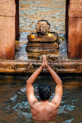A person is standing waist-deep in water with their hands together in a prayer gesture. In front of them is a small stone sculpture with intricate carvings, adorned with flowers. The scene appears to be part of a ritual or prayer, with the water and stone structure playing significant roles.