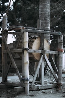 A rustic wooden structure with bamboo supports stands on a dirt ground. The construction appears to be handmade, possibly for a practical purpose. There are ropes tied around the bamboo poles, and the background includes a large tree trunk and greenery.