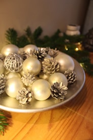 A decorative plate filled with silver pine cones and white ornaments, placed on a wooden table. The background includes green pine branches and small lights, creating a festive ambiance.