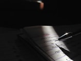 A close-up view of a pen resting on a notebook, with handwritten text visible on the pages. The scene is partially illuminated by a focused light source, creating dramatic shadows and emphasizing the written content.