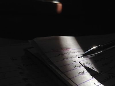 A close-up view of a pen resting on a notebook, with handwritten text visible on the pages. The scene is partially illuminated by a focused light source, creating dramatic shadows and emphasizing the written content.