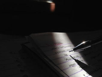 A close-up view of a pen resting on a notebook, with handwritten text visible on the pages. The scene is partially illuminated by a focused light source, creating dramatic shadows and emphasizing the written content.