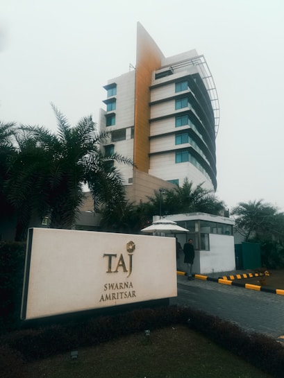 A modern multi-story hotel building with a distinctive architectural design featuring curved lines and a mix of beige and blue tones. The foreground shows a large sign with the name 'Taj Swarna Amritsar', partially surrounded by neatly manicured greenery.
