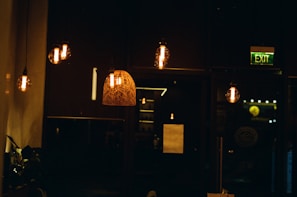 Dimly lit interior setting with hanging light bulbs and a decorative lamp. The lights cast a warm glow, creating a cozy ambiance. There is a visible exit sign in green and some plants partially visible on the left.
