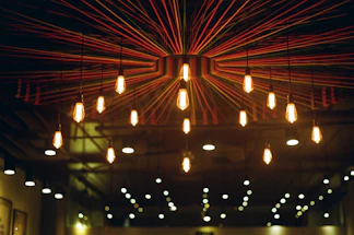 A ceiling installation featuring hanging light bulbs with a radial design of colorful strings extending from a central point. The warm glow from the bulbs creates a cozy and inviting atmosphere.