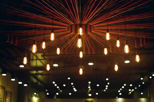 A ceiling installation featuring hanging light bulbs with a radial design of colorful strings extending from a central point. The warm glow from the bulbs creates a cozy and inviting atmosphere.
