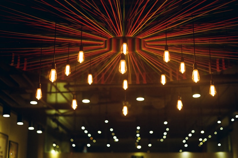 A ceiling installation featuring hanging light bulbs with a radial design of colorful strings extending from a central point. The warm glow from the bulbs creates a cozy and inviting atmosphere.