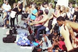 A vibrant street performance scene with musicians playing drums while a diverse crowd watches. People of various ages and backgrounds gather, some clapping and others simply observing. The atmosphere is lively, with a pile of colorful bags and backpacks nearby, suggesting a casual, outdoor setting.