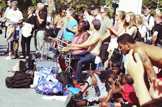 A vibrant street performance scene with musicians playing drums while a diverse crowd watches. People of various ages and backgrounds gather, some clapping and others simply observing. The atmosphere is lively, with a pile of colorful bags and backpacks nearby, suggesting a casual, outdoor setting.