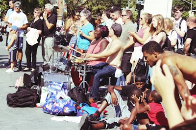 A vibrant street performance scene with musicians playing drums while a diverse crowd watches. People of various ages and backgrounds gather, some clapping and others simply observing. The atmosphere is lively, with a pile of colorful bags and backpacks nearby, suggesting a casual, outdoor setting.