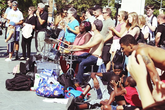 A vibrant street performance scene with musicians playing drums while a diverse crowd watches. People of various ages and backgrounds gather, some clapping and others simply observing. The atmosphere is lively, with a pile of colorful bags and backpacks nearby, suggesting a casual, outdoor setting.