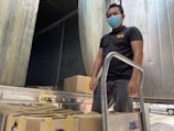 A person wearing a blue mask and black shirt stands beside a hand truck loaded with sealed cardboard boxes. The background features a metal-lined container or truck with open doors.
