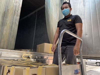 A person wearing a blue mask and black shirt stands beside a hand truck loaded with sealed cardboard boxes. The background features a metal-lined container or truck with open doors.