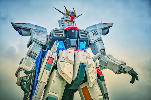 A large, detailed robot model stands against a cloudy sky. The design features intricate mechanical parts, with prominent sections in white, blue, red, and gray. The robot has a humanoid structure with a head, arms, and legs, and its head is adorned with sharp, angular elements and antennae.