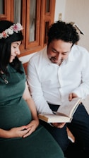 A couple is sitting together, with the woman wearing a floral headband and a green dress gently touching her pregnant belly. The man is smiling while holding an open book. They are in an indoor setting near a wooden window.
