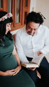 A couple is sitting together, with the woman wearing a floral headband and a green dress gently touching her pregnant belly. The man is smiling while holding an open book. They are in an indoor setting near a wooden window.