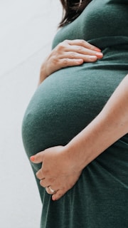 A person gently cradles their pregnant belly with both hands, showcasing a close-up view of the rounded shape. The person is wearing a dark green dress, and the focus is on the lower torso with one hand positioned above and the other below the belly.