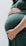 A person gently cradles their pregnant belly with both hands, showcasing a close-up view of the rounded shape. The person is wearing a dark green dress, and the focus is on the lower torso with one hand positioned above and the other below the belly.