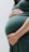 A person gently cradles their pregnant belly with both hands, showcasing a close-up view of the rounded shape. The person is wearing a dark green dress, and the focus is on the lower torso with one hand positioned above and the other below the belly.