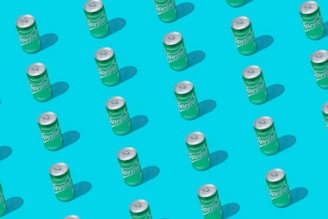 Multiple green cans organized in a neat, diagonal pattern on a vibrant blue background. Each can casts a shadow, enhancing the repetitive design and creating a sense of depth.
