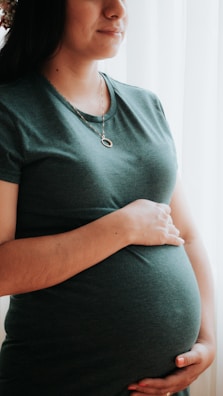A woman wearing a green t-shirt is standing by a window, gently holding her pregnant belly. She wears a simple necklace and looks content and serene.