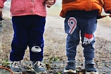 Two young children stand side by side outdoors. They are dressed in winter clothing, with one wearing a pink sweater and dark blue pants featuring a bear face, and the other in an orange jacket and grey pants with a number nine design. They both have sneakers on, surrounded by fallen leaves on the ground.