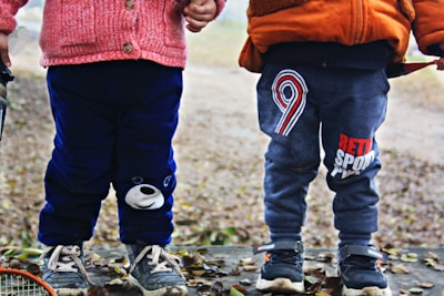 Two young children stand side by side outdoors. They are dressed in winter clothing, with one wearing a pink sweater and dark blue pants featuring a bear face, and the other in an orange jacket and grey pants with a number nine design. They both have sneakers on, surrounded by fallen leaves on the ground.