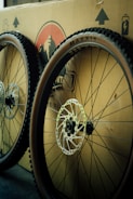 Two bicycle wheels with knobby tires are leaning against a cardboard box. The wheels feature disc brakes, and the box has printed graphics and symbols on it, including a logo and arrows indicating direction.