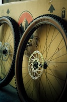 Two bicycle wheels with knobby tires are leaning against a cardboard box. The wheels feature disc brakes, and the box has printed graphics and symbols on it, including a logo and arrows indicating direction.