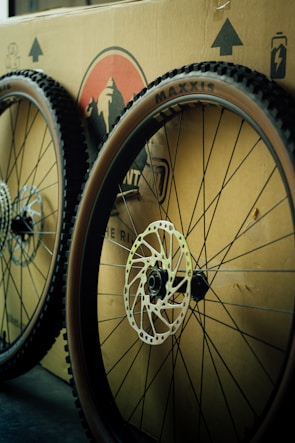 Two bicycle wheels with knobby tires are leaning against a cardboard box. The wheels feature disc brakes, and the box has printed graphics and symbols on it, including a logo and arrows indicating direction.