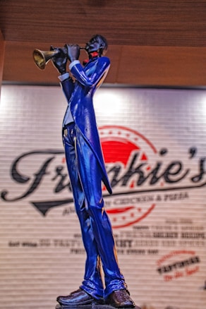 A statue of a musician playing a trumpet, wearing a striking blue suit and standing in front of a wall with a logo that appears to be for a restaurant or caf&eacute;.