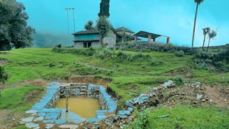 A rural landscape with vibrant green grass, rustic stone structures, and a small body of water surrounded by stones. A traditional house with colorful prayer flags and a sloping roof sits atop a hill. Sparse trees and electric poles are visible under a clear sky.