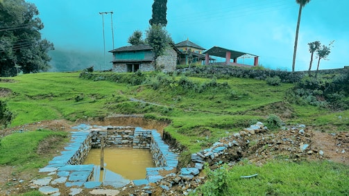 A rural landscape with vibrant green grass, rustic stone structures, and a small body of water surrounded by stones. A traditional house with colorful prayer flags and a sloping roof sits atop a hill. Sparse trees and electric poles are visible under a clear sky.
