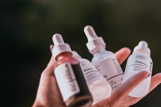 A hand holding several dropper bottles with labels, likely skincare products. The bottles vary in color, including shades of white, pink, and clear, with branding visible on the labels.
