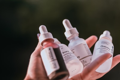 A hand holding several dropper bottles with labels, likely skincare products. The bottles vary in color, including shades of white, pink, and clear, with branding visible on the labels.