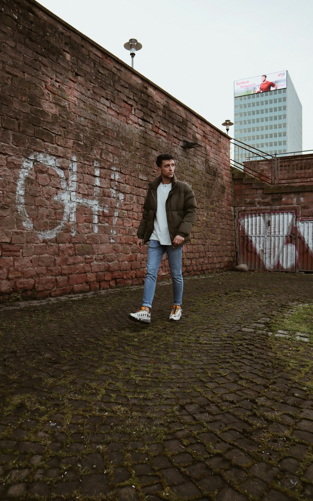 man in gray jacket and blue denim jeans standing beside brick wall