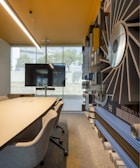 A modern conference room with a large meeting table and several chairs on wheels. The room features a wall adorned with abstract art, which includes various shapes and patterns. A large flat-screen television is mounted on a stand at the end of the table, set against a window that allows natural light to flood the space. The ceiling has a perforated design with recessed lighting, and the overall decor is sleek and contemporary.