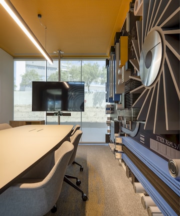 A modern conference room with a large meeting table and several chairs on wheels. The room features a wall adorned with abstract art, which includes various shapes and patterns. A large flat-screen television is mounted on a stand at the end of the table, set against a window that allows natural light to flood the space. The ceiling has a perforated design with recessed lighting, and the overall decor is sleek and contemporary.