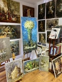 A room filled with various paintings displayed on easels and mounted on the walls. The largest painting in the center shows a bouquet of white flowers against a blue background. Surrounding it are several other artworks featuring different subjects like a vintage teapot, abstract figures, and landscapes. On the right side, there is a wooden bookshelf with books and more framed artwork.