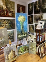 A room filled with various paintings displayed on easels and mounted on the walls. The largest painting in the center shows a bouquet of white flowers against a blue background. Surrounding it are several other artworks featuring different subjects like a vintage teapot, abstract figures, and landscapes. On the right side, there is a wooden bookshelf with books and more framed artwork.
