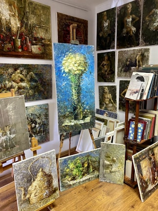 A room filled with various paintings displayed on easels and mounted on the walls. The largest painting in the center shows a bouquet of white flowers against a blue background. Surrounding it are several other artworks featuring different subjects like a vintage teapot, abstract figures, and landscapes. On the right side, there is a wooden bookshelf with books and more framed artwork.