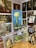 A room filled with various paintings displayed on easels and mounted on the walls. The largest painting in the center shows a bouquet of white flowers against a blue background. Surrounding it are several other artworks featuring different subjects like a vintage teapot, abstract figures, and landscapes. On the right side, there is a wooden bookshelf with books and more framed artwork.