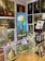 A room filled with various paintings displayed on easels and mounted on the walls. The largest painting in the center shows a bouquet of white flowers against a blue background. Surrounding it are several other artworks featuring different subjects like a vintage teapot, abstract figures, and landscapes. On the right side, there is a wooden bookshelf with books and more framed artwork.