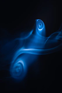 Swirls of neon blue light create ethereal patterns against a dark background, resembling smoke or liquid in motion. The forms suggest a sense of fluidity and mystery.