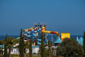 A series of colorful water slides with multiple twists and turns is set against a backdrop of blue ocean under a clear sky. Surrounding the slides, there are various green trees and plants, adding a natural element to the scene.