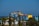 A series of colorful water slides with multiple twists and turns is set against a backdrop of blue ocean under a clear sky. Surrounding the slides, there are various green trees and plants, adding a natural element to the scene.