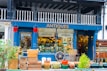 A charming antique shop display featuring a variety of vintage items. The exterior is painted dark blue with the word 'ANTIQUES' prominently displayed above the entrance. The storefront is filled with various antique objects, including wooden crates, earthen pots, a ship's wheel, and a stack of vintage suitcases. Red fire buckets hang on either side of the entrance. The interior is bustling with a diverse collection of antiques, adding to the shop's allure.