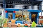 A charming antique shop display featuring a variety of vintage items. The exterior is painted dark blue with the word 'ANTIQUES' prominently displayed above the entrance. The storefront is filled with various antique objects, including wooden crates, earthen pots, a ship's wheel, and a stack of vintage suitcases. Red fire buckets hang on either side of the entrance. The interior is bustling with a diverse collection of antiques, adding to the shop's allure.